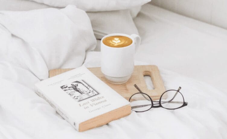 What Self Care Really Means - A coffee, a book, and reading glasses over a bed.