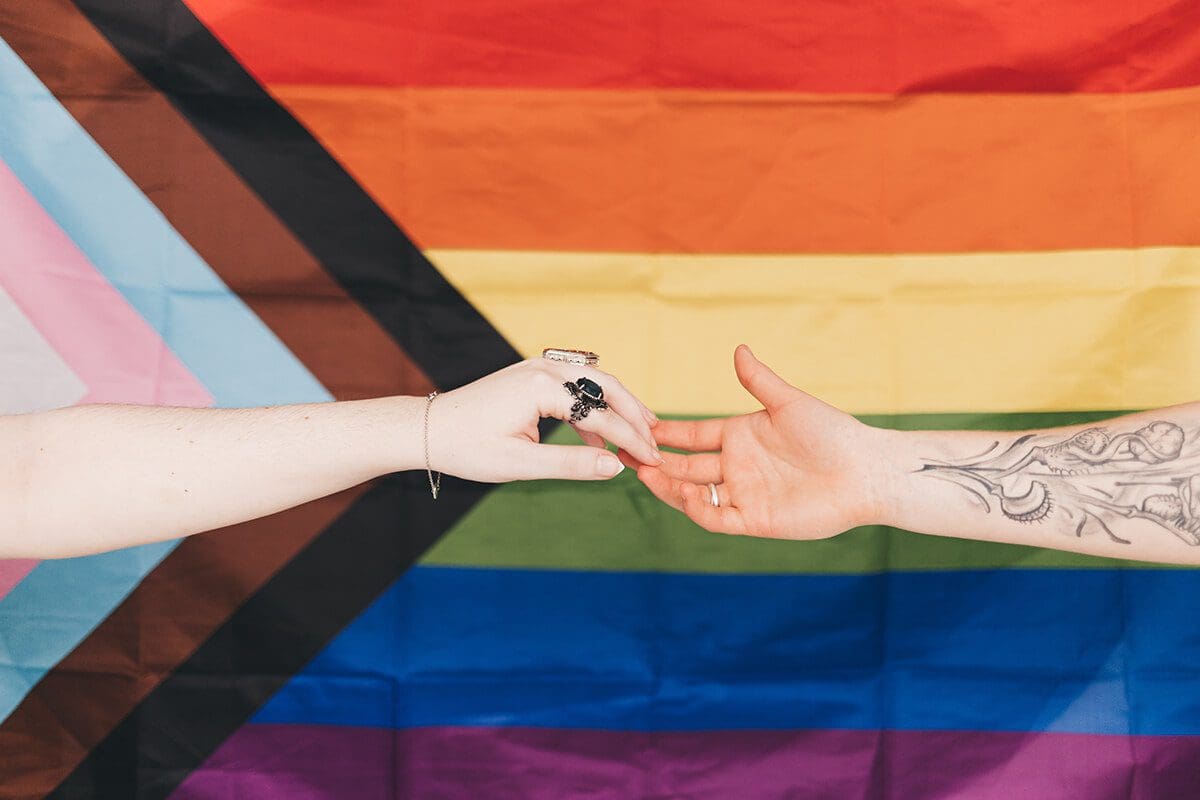 Guidely supports the LGBTQ+ Community. Image of two hands reaching out to each other, in front of the Progress Pride flag.