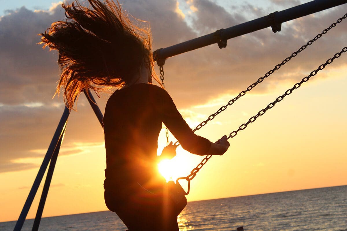 10 amazing ways to reach happiness in life and work. Image shows a woman on a swing, with a sunset behind.